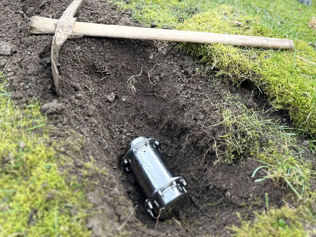 A metal time capsule lies in a freshly dug hole, with a tool beside it.